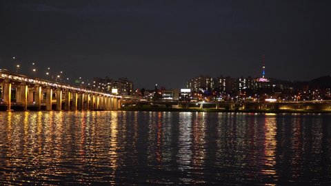 video at night of the Han River, Seoul South Korea, the iconic Banpo bridge and Namsan Tower. The foreground features beautiful colored reflections of city lights on the water