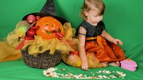 A little girl in a witch costume sits on a green background sprinkled with beans, walnuts and pumpkin seeds, collects in a pink baby pot