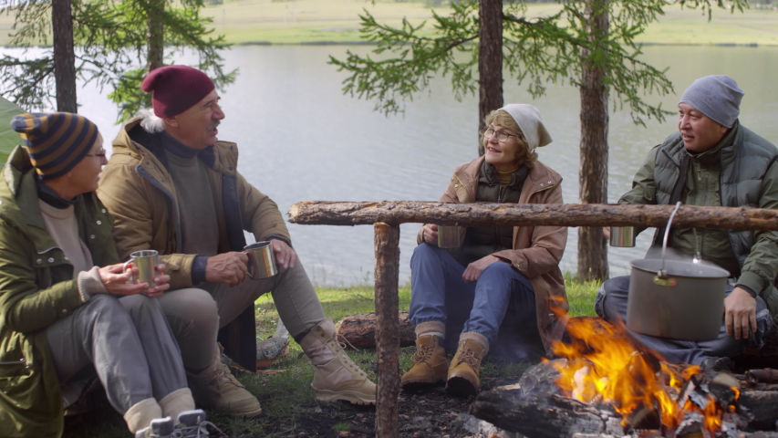 Cheerful senior men and women smiling and discussing something while sitting together beside fireplace at campsite and having tea