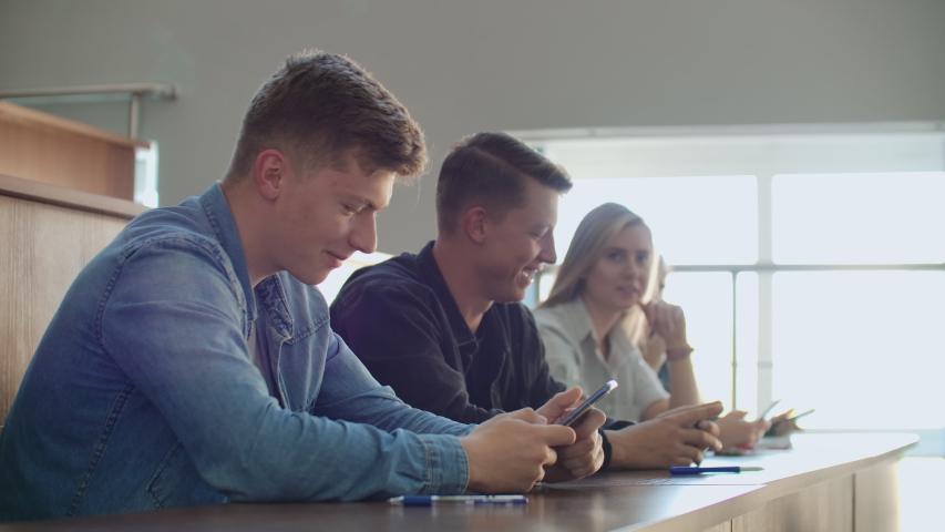 Two friends are sitting in a large classroom with students and showing each other funny videos from the boarding school.