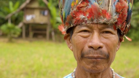 portrait of an old indigenous amazonian man standing in his village wearing traditional outfit and a colorful hat slow- motion closeup shot in ecuador