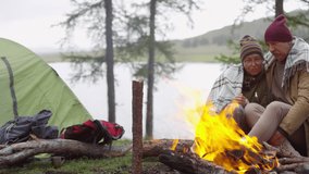 Tracking shot of romantic senior couple sitting on wooden log under blanket, looking at burning campfire, embracing and discussing something - Powered by Shutterstock - Get 15% off with code: PIKWIZARD15