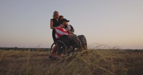 Full shot of a wheelchaired American soldier watching sunset with his family on a meadow