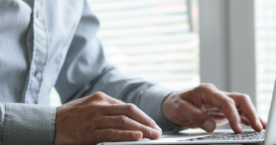 man on a stilysh shirt typing at his laptop computer in an office next to the window