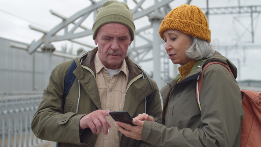 Pan shot of Caucasian elderly man and woman standing on train station, using smartphone and smart watch and talking