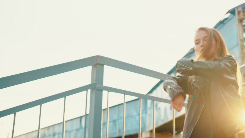 Young girl teenager dancing front camera on staircase on sunshine background. Rock girl in black leather jacket dancing on urban stairs at bright sunset landscape