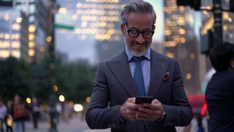 Slow motion of smiling mature lawyer in expensive suit messaging with customers via cellular device while walking in financial district,successful businessman in optical eyewear searching trading news
