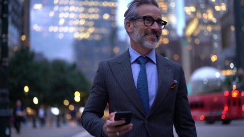 Successful male entrepreneur in elegant suit walking at metropolitan street in downtown and smiling during online messaging via smartphone device,slow motion effect of confident businessman in eyewear