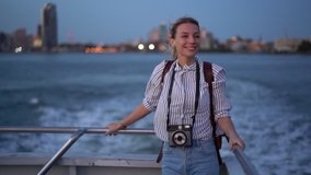 Smiling female blogger in casual clothing using cellphone camera for clicking pictures of evening metropolitan cityscape during water boat excursion, young hipster girl photographing via cellular
