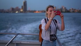 Happy female blogger in casual clothing using cellphone camera for clicking pictures of evening metropolitan cityscape during water boat excursion, cheerful hipster girl photographing via cellular
