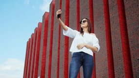 Beautiful woman tourist uses a mobile phone for video call to friends near a modern brick building, exhibition center, communication concept