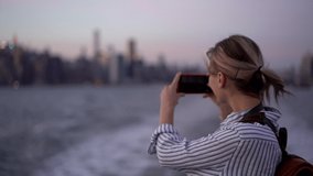 Slow motion effect of millennial woman tourist photographing cityscape during water excursion using modern smartphone camera, happy Caucasian female enjoying voyage trip during evening time