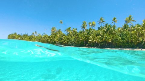 HALF UNDERWATER, SLOW MOTION: Spectacular turquoise water surrounds the exotic sandy beach of One Foot Island in the Pacific. Idyllic view of the glassy ocean and the white sand coast with palm trees