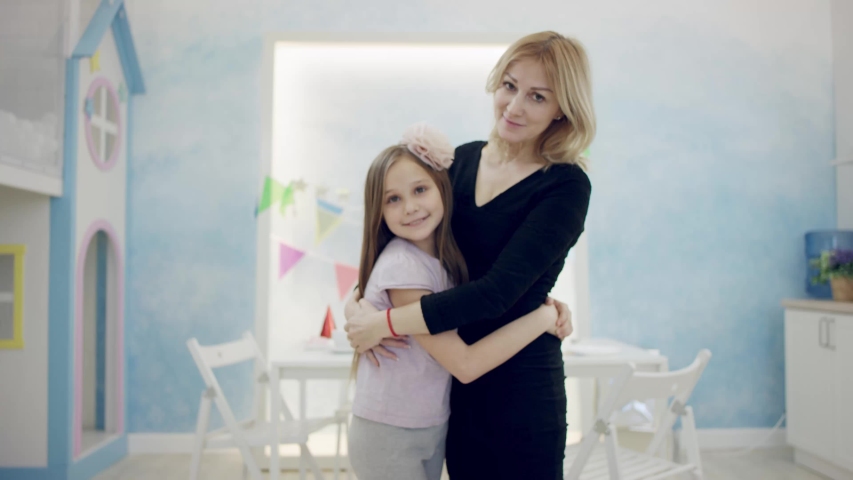 Adult woman is hugging and kissing her little daughter in front of the camera in a room