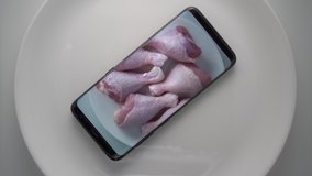 Cell phone on plate with fresh raw chicken drumsticks on screen in microwave. Food blog concept. Top view of mobile phone with food footage on screen rotating in microwave oven.