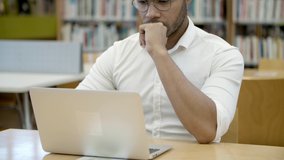 Focused young man working with laptop in library. Thoughtful African American student in eyeglasses sitting at table and typing on laptop. Education concept