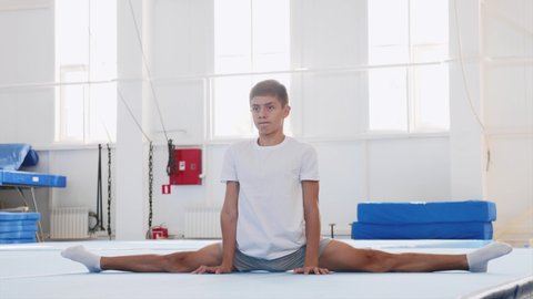 A teenager boy gymnast in white T-shirt is making a front split in the sport center in front of the camera. Acrobatic equipment at the background.
