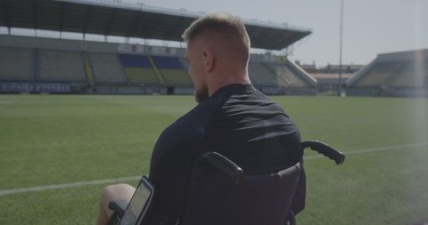 Medium shot of a sad disabled athlete sitting in his wheel chair by a sports field and looking away