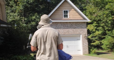 A worker on a landscape crew walks across a yard and driveway carrying a blue tarp in his hands on a hot summer day