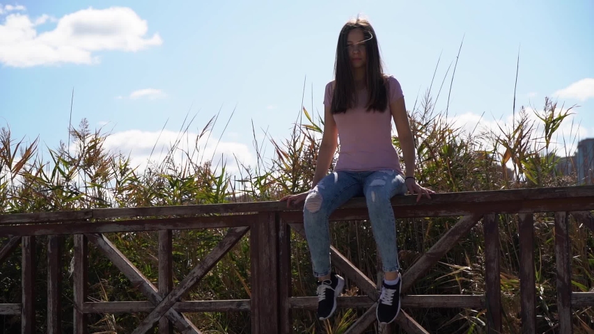 Nice attractive girl in pink t-shirt and ripped blue jeans sit on rails and fix her hair. Alone girl self isolation quarantine coronavirus.