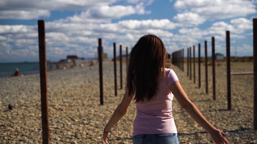 Pretty, attractive girl or young woman in pink shirt and blue ripped jeans spinning around herself on the beach with nice blue cloudy sky on background. Slow motion
