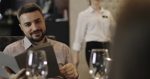 The waiter came to the customers. Portrait of bearded man sitting at the table in restaurant and readding menu. Man is talking, smiling, laughing and making order food and drinks.