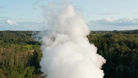 Aerial drone shot of a factory chimney spewing fumes into atmosphere. Fly through video of a thick smoke plume rising in the blue sky. Forest and meadow in the background. Summer day. 4k footage.