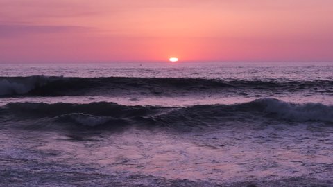 Waves in slow motion at sunset. Beautiful pastel pink / orange sky, Sun on horizon in distance.