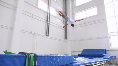 A teenage athlete is jumping on the sport trampoline in the gym and in slow motion does a back flip in the air. Lands on feet with hands up. Green and blue sport slings in the foreground. Side view.