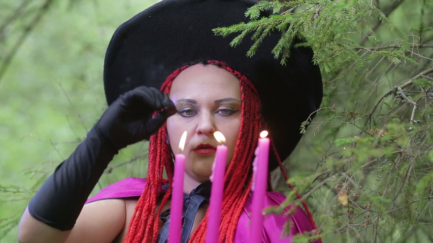 young witch with red hair and a pointed hat and a raspberry cloak conjures with candles in the forest