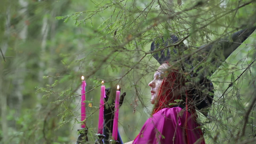 a young witch with red hair and a black hat and a raspberry cloak practices magic in the forest.