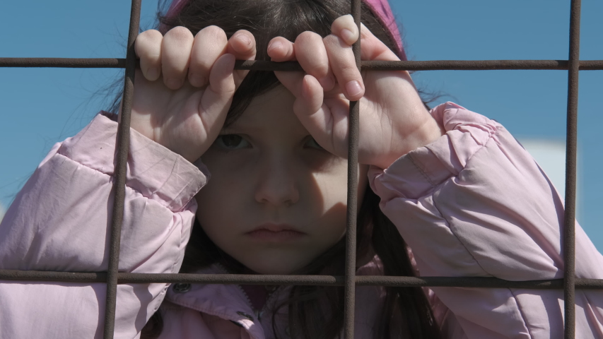 Sad child behind the fence. Homeless little girl behind an iron fence. Child in a cage.