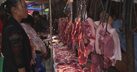 Chengdu.Sichuan/China-September 27th 2019: people shopping pork in the fresh market, old style open outdoor food market street young female vendor busy on her stall red meat hanging in sunshine