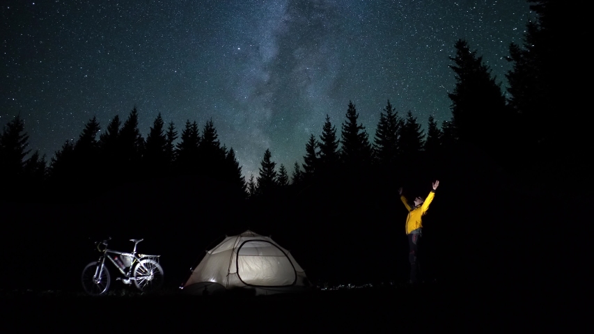 A man looks at the stars near the tent in the night forest. Star trail move in the night sky. Timelapse. Travel concept. 4K