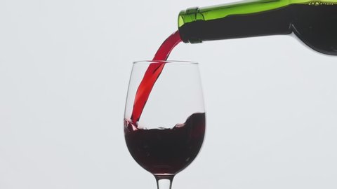 Red wine pouring in wine glass at white background. Close-up shot. Slow motion of pouring red wine from bottle into goblet. Red wine forms beautiful wave in 4k, UHD