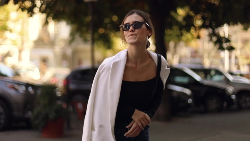 Beautiful woman looks at the clock waiting for someone. Hurry up! A woman nervously walks from side to side waiting for someone else. Girl wear white blazer, black sunglasses and black top. 