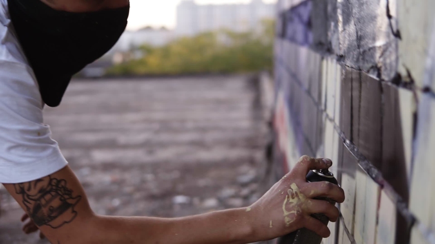 Close-up of artists hand dirty in paint applying spray drawing a black colour on a street building wall. Action. Stained fingers of artist hold spray can with colored paint on concrete wall. Street