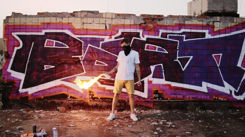 Scene of a graffiti urban artist in balaclava after painting the his picture on the street wall with spray paints. Standing against the painted wall with fire stream burning from the spray can