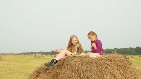 Cute girls sitting on haystack. Vertical motion of two adorable girls in checkered shirts sitting on haystack and talking in field during harvest.