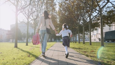 Young woman and little girl in school uniform are holding hands and running along the trees in the park at sunny autumn weather. Family late for school concept, steadicam shot back view.