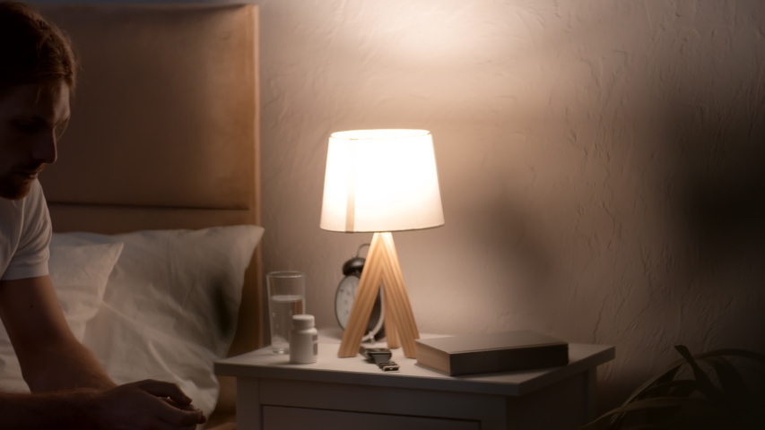 Tracking shot of handsome young man in t-shirt sitting on his bed and taking sleeping pills, then getting under covers and turning off lamp on nightstand