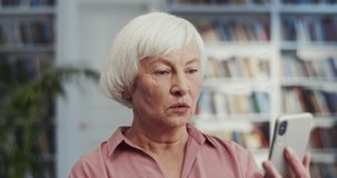 Old Caucasian grey-haired woman holding a smartphone, looking at the screen and seeing or reading something scary and awful. Close up.