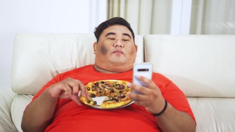 Overweight man eating a plate of pizza while using mobile phone on the sofa in the living room at home. Shot in 4k resolution
