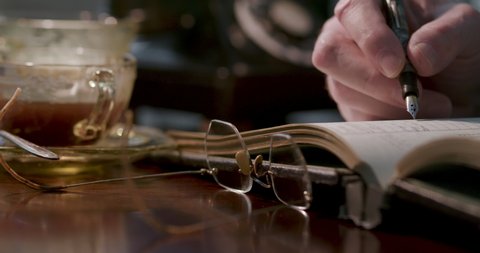 Scene tracks from depression period glass coffee cup to the hand of a man writing in a ledger with a classic ink fountain pen and vintage eyeglasses laying on the desk in front of him.