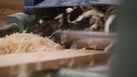 A solid wood plank coming out from an old planing machine. Slow motion shot of the plane tool processing wood while wood shavings are flying towards the camera. Shot taken in a woodworking workshop.
