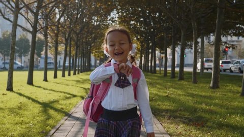 Child girl in school uniform is walking quickly in the park and talking on her pink wrist smart watches and laughing at sunny autumn weather, trees along the way, front view, steadicam shot.