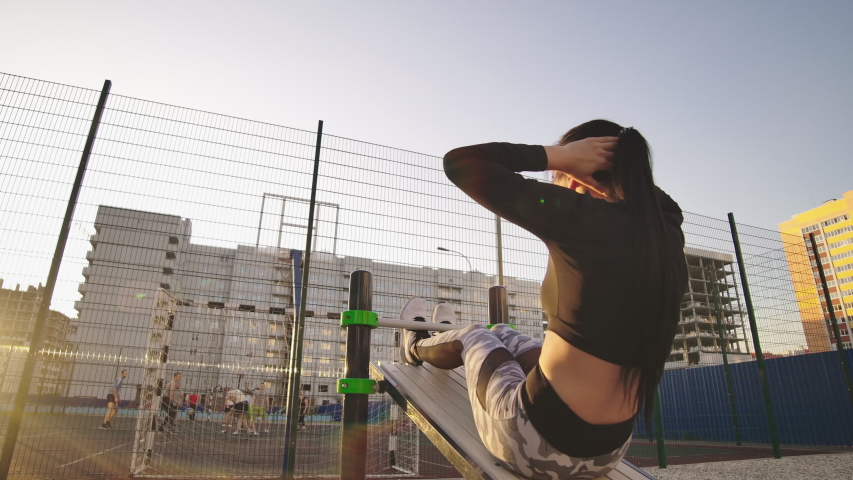 Doing twisting crunches. Serious confident sporty young woman sitting on wooden bench and doing twisting crunches at sports ground.