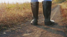 Close-up of a farmer's feet in rubber boots walking down a country road between fields, dust rising from shoes. Slow-motion 4k video