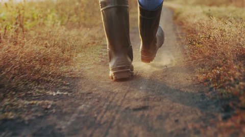 Close-up of a farmer's feet in rubber boots walking down a country road between fields, dust rising from shoes. Slow-motion 4k video