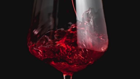 Red wine forms beautiful wave. Wine pouring in wine glass over black background. Close-up shot. Slow motion of pouring red wine from bottle into goblet. Low key